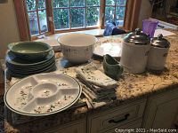 Overview of the lot showing the canisters, plates, bowls, Christmas placemats, napkins, and purple glass vase on kitchen counter