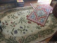 Photo displaying large oval beige floral rug with green leaf and multi-color flower border, plus two smaller rugs stacked on top