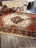 Full view of one large rug partially folded on a wood floor, showing intricate floral and geometric patterns in brown, cream, blue-gray, and rust-orange tones.