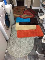 Photo showing two oval braided rugs in green and multicolor tones and assorted mats in different colors and shapes on tiled floor near washer.