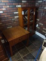 Photo of wood side table and wood cabinet with glass paneled doors set against a brick wall on tile floor.