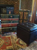 Photo showing the book-themed cabinet and side table with painted book spines and the decorative snowman box on top, along with the dark brown leather storage ottoman.