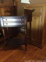 Photo showing the dark wood side table with brass drawer knobs, turned legs on caster wheels, and lower shelf, positioned next to the lighter wood side table.