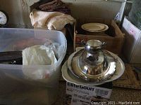 View of slow cooker box with silver plate serving dish and other assorted dishes in boxes and plastic bin containing kitchen linens and some utensils.
