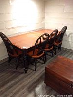 Photo showing the wood dining table with four matching chairs arranged in a corner. Warm medium brown finish on table, darker stained chair legs and spindled backs. Table top shows signs of wear.