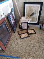 Overview of all framed art pieces placed on floor, including cross-stitch samplers, patriotic eagle print, and owl print, showing condition and size relative to floor.