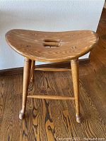 Full view of the stool showing light oak wood, saddle-shaped seat with cutouts, turned legs, and stretchers on wood floor.