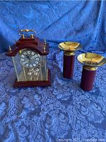 Anniversary clock and two crimson brass candle holders together on blue patterned cloth