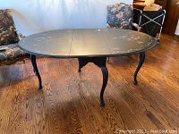 Oval gate leg dining table painted black with hand painted white floral designs on the surface and curved legs.