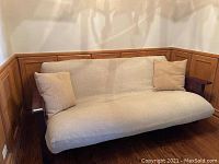 Photo of futon showing full couch form with light beige upholstered mattress and two beige pillows on either side, solid cherry wood frame with armrests.