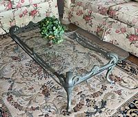 Rectangular coffee table with patina bronze metal base and ornate detailing, topped by beveled removable glass, sitting on a floral rug between floral-patterned couches.