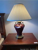Full view of the maroon and brass table lamp on a wooden surface with pleated beige lampshade illuminated.