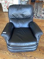 Front view of black leather padded glider recliner chair, showing cushioned armrests, backrest, and seat with some creasing on leather surface.