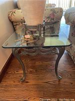 Full view of the square patina bronze metal base end table with a removable beveled edge glass top, situated between floral patterned upholstered furniture on wooden flooring.