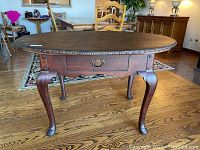 Full view of the oval mahogany library table with leather top and single center drawer featuring brass-like pull, standing on sculptured cabriolet legs with dark walnut finish.