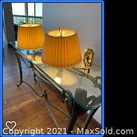 Two brass wall lamps with off-white pleated shades positioned on a glass table, showing reflective view and light on.