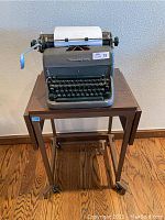 Front view of the Remington Rand typewriter on the Tiffany rolling stand on hardwood floor, against plain wall.