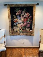 Full view of the floral still life tapestry hung on a wall between two chairs, showing the bronze rod and decorative finials included for mounting.