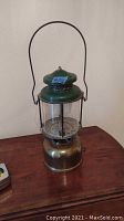 Full view of antique Coleman lantern showing brass base, green painted top with vent holes, clear glass globe, and large arched handle.