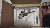 Bird figurine with red head, blue tail, green body and white flower next to clear glass stand inside a box with measuring tape showing about 12 inches