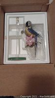 Blue parrot figurine and glass stand packaged in styrofoam and plastic inside box.