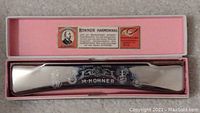Metal harmonica with M. Hohner engraved, inside a pink presentation box with a label and text about the brand and maker.