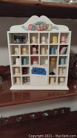 Front view of white wooden shelving unit filled with various sewing thimbles in compartments. Shelf has sewing-themed decorations at top.