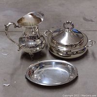 Photo showing silver pitcher, covered casserole dish, and oval platter on concrete floor.