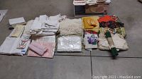 Photo of a spread of various folded linens including tablecloths, napkins, tea towels, crochet pieces, and pillowcases arranged on a floor surface.