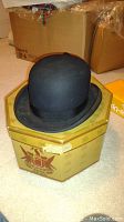 Black bowler hat resting on an octagonal hat box with crease and decorative graphics.