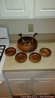Overall view of large wooden salad bowl, five smaller serving bowls, and pair of wooden salad utensils on kitchen countertop.