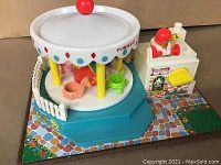 Side view of vintage Fisher Price Musical Carousel toy showing white carousel top with colorful decorations, animal figures on platform, and adjacent ticket booth with red hat figure and on/off switch.