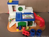 Front view of the Fisher Price Sesame Street Clubhouse showing the white clubhouse with blue roof, green door, red slide, orange base, and four colorful peg figures and toy accessories in front.