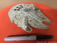 Front angled view of white diecast Millennium Falcon with grey and brown spots, next to a black Sharpie for size reference.