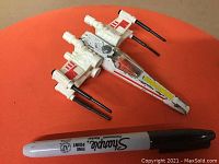 Image of the entire X-Wing Fighter toy on an orange surface alongside a black Sharpie pen for size comparison.