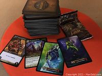 Stack of about 250 World of Warcraft trading cards on an orange surface, along with a few individual cards showing game character and item artwork and one unopened booster pack.