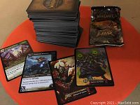 Stack of approximately 250 World of Warcraft trading cards upright, alongside a few cards laid out face-up showing character and creature artwork, plus one unopened booster pack.