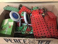 Box holding assorted early Meccano parts including perforated metal plates in red and green, wheels, and metal rods.
