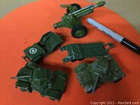 Photo showing five green military toy vehicles on an orange background with a black marker for scale. Vehicles include an artillery cannon, tanks, a Jeep style vehicle, and a flatbed trailer.