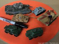 View of the collection of seven vintage military toy tanks and armored vehicles arranged around a Sharpie pen for scale, showing tops and sides of models.