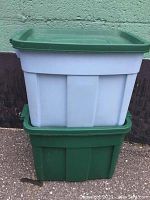 Two large Rubbermaid Roughneck storage bins stacked outdoors on pavement against a green wall, showing a green bin with matching green lid on bottom and a gray bin with green lid on top.