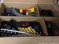 Two cardboard boxes filled with plastic slot car track sections, two wired controllers, railings, and two slot cars (one black and one red).