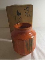Ceramic octagonal vase with orange-red glaze and green vertical streaks, shown with its wooden box marked with Chinese characters.