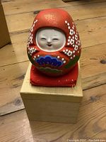 Front view of the Japanese clay Daruma figurine showing the smiling white face and red floral-painted body sitting on a red cushion and wooden box.