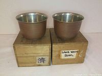 Side view of the two matching metal sake waste bowls sitting on top of their original wooden boxes labeled with Japanese characters and English text.
