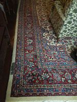 Photo showing a corner section of the carpet with detailed floral borders in reds and blues against cream background, next to furniture.