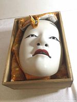 Front view of the painted ceramic Noh mask inside its original wooden box lined with orange fabric.