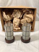 Two capriz shell votive covers standing next to a box with packing paper inside, showing multiple units.