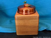Front view of a square wooden storage box with copper lid topped by a small wooden duck figurine.