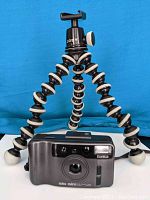 Photo showing Joby GorillaPod tripod with its flexible segmented legs and Konica Big Mini film camera in front of it, against a blue background.